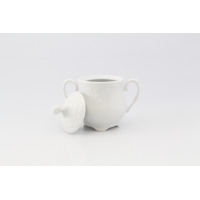 Biała porcelanowa cukiernica 0,25 l z serii Rococo marki Ćmielów. Klasyczne naczynie z ozdobnymi tłoczeniami, dwoma uchwytami i zdjętą pokrywką na jasnym tle.