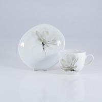 Biała porcelanowa filiżanka 0,20 l ze spodkiem Lubiana 6474 Magnolia. Zestaw zdobi subtelny, szary motyw kwiatowy. Widok z boku na tle o neutralnej barwie.