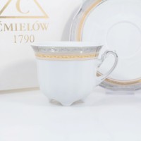 Biała porcelanowa filiżanka 0,23 l ze spodkiem Ćmielów E551 Bolero, zdobiona złotym i srebrnym ornamentem. W tle widoczne logo marki Ćmielów 1790.