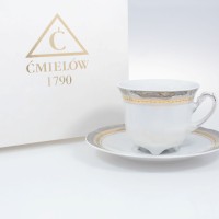 Biała porcelanowa filiżanka ze spodkiem Ćmielów E551 Bolero o pojemności 0,23 l. Zdobiona złotym ornamentem, stoi obok pudełka z logo marki i napisem ĆMIELÓW