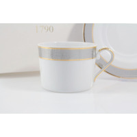 Biała porcelanowa filiżanka Ćmielów E877 Jenny o pojemności 0,25 l z eleganckim srebrno-złotym zdobieniem przy krawędzi. W tle widoczny spodek oraz napis 1790.
