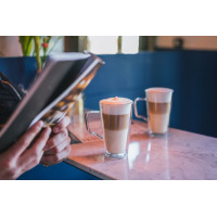 Dwie szklanki do latte z podwójną ścianką Vialli Design DIVA wypełnione warstwową kawą na marmurowym blacie, obok osoba czytająca książkę w kawiarnianym
