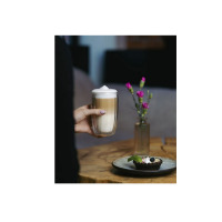 Dłoń trzyma wysoką szklankę termiczną 350 ml TULIP od Vialli Design wypełnioną warstwowym latte. Obok na drewnianym stole stoi wazon z kwiatami i deser na