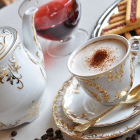 Elegancki garnitur do kawy Ćmielów 7830 ROCOCO Anna. Biała porcelana ze złotymi zdobieniami, filiżanka z cappuccino, dzbanek, ciasteczka i ziarna kawy na stole.