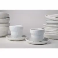 Dwie perłowe filiżanki Verlo Rustic Reborn 160 ml na spodkach, ustawione na tle stosów ceramicznych naczyń. Minimalistyczny design, jasna kolorystyka.