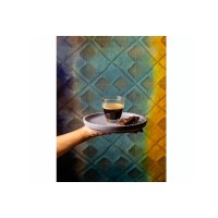 Dłoń trzymająca szklankę do espresso Enigma marki Onis o pojemności 110 ml z kawą, serwowaną na liliowym talerzyku z kawałkiem czekolady na tle wzorzystej
