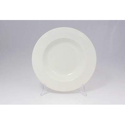 Talerz do pasty 30 cm BANQUET marki RAK Porcelain w kolorze kremowej bieli. Okrągłe naczynie z szerokim rantem ustawione na przezroczystym stojaku, widok od
