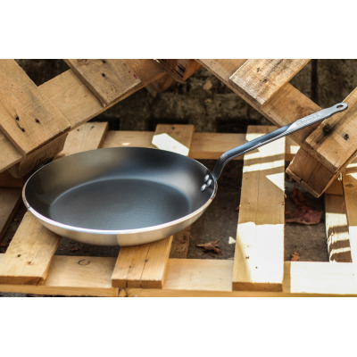 Patelnia 32 cm CHOC Resto Induction marki De Buyer z nieprzywierającą powłoką, ustawiona na drewnianych paletach. Widoczny napis na rączce: de Buyer MADE IN
