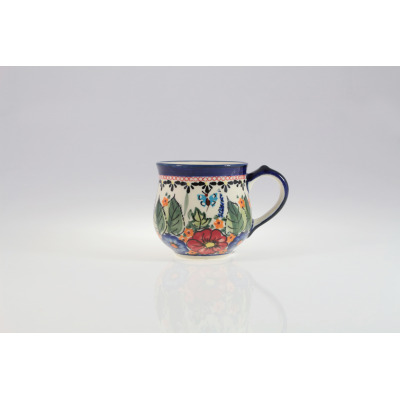 Ceramiczny kubek Bolesławiec 0,23 l GU-1472 z ręcznie malowanym motywem kwiatów i błękitnego motyla na kremowym tle, ujęty na wprost na białym tle z odbiciem.