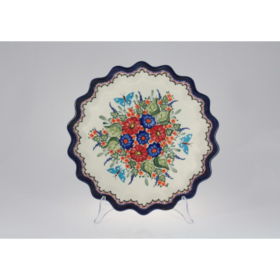 Ceramiczny talerz z falbanką Bolesławiec GU-1332A o średnicy 32,4 cm. Kremowe tło zdobi ręcznie malowany wzór w czerwone i niebieskie kwiaty, zielone liście i